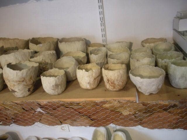 Multiples bols ou pots artisanaux en papier mâché ou matière similaire, aux bords irréguliers, alignés sur une étagère en bois, devant un mur blanc.