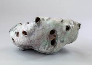 Sculpture céramique organique, bosselée, à glaçure craquelée blanc-vert-rose et percée de trous sombres.