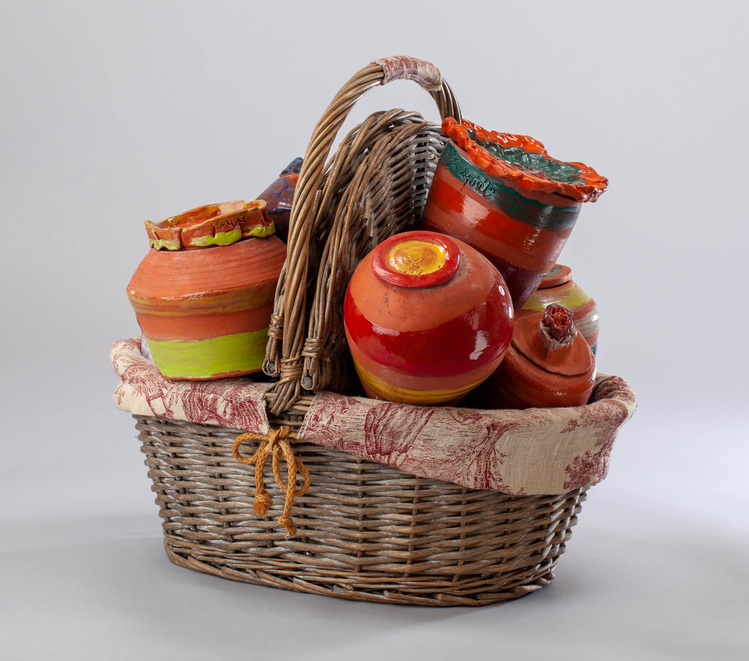 Panier en osier rempli de poteries en terre cuite colorées, rouges, jaunes et vertes, doublé d'un tissu à motifs toile de Jouy.