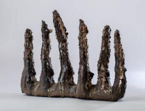Sculpture céramique abstraite brune et noire texturée, rappelant des dents ou des stalagmites fondues, sur fond gris clair.