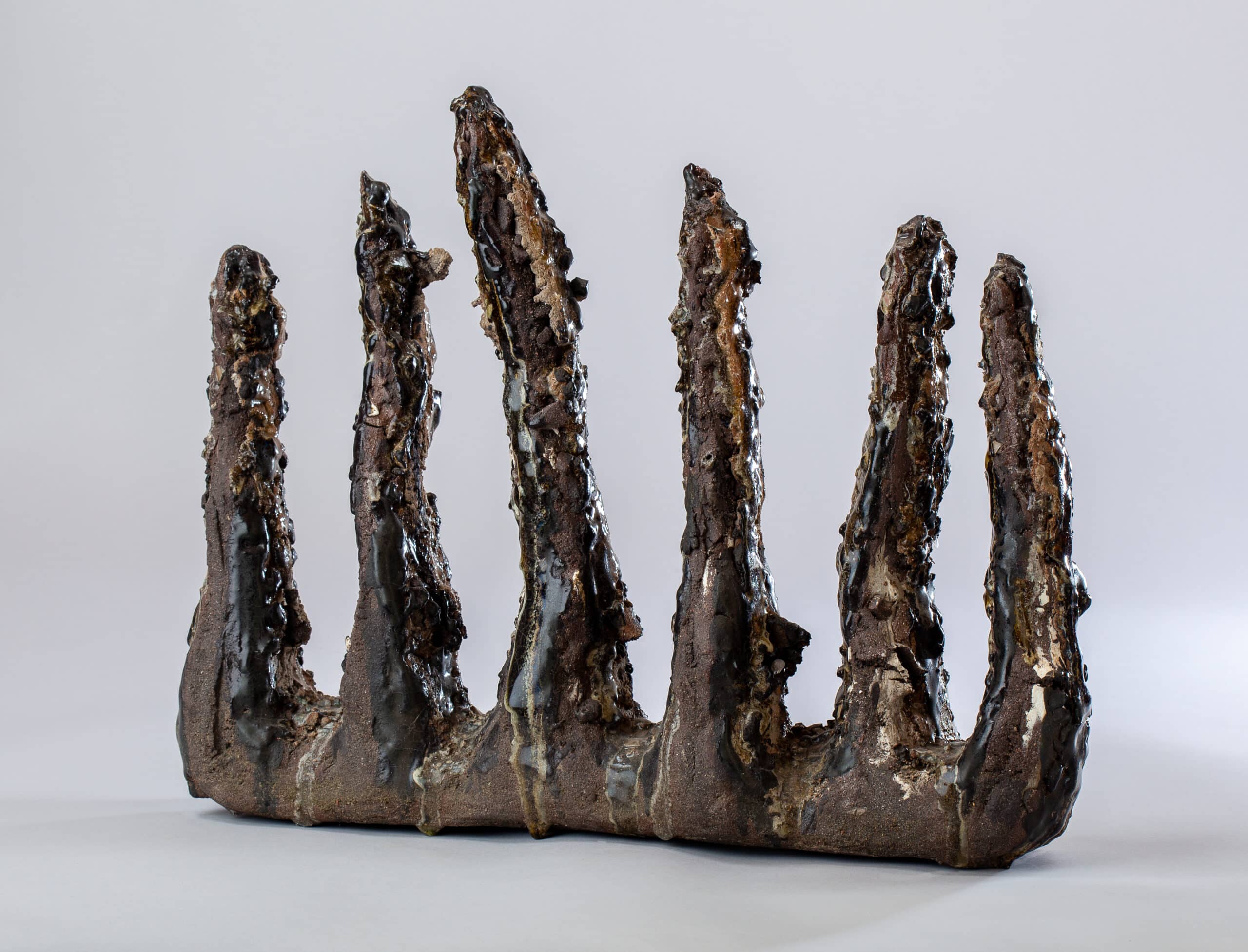 Sculpture céramique abstraite brune et noire texturée, rappelant des dents ou des stalagmites fondues, sur fond gris clair.