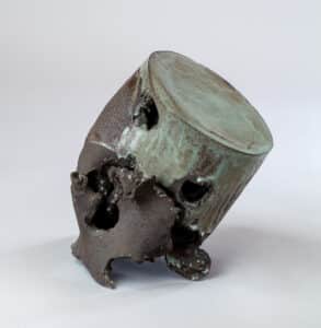 Sculpture céramique abstraite inclinée, texture brute noire et glaçure bleu-vert patinée avec ouvertures irrégulières.