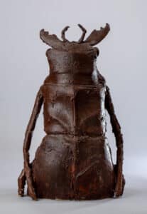 Sculpture abstraite d'un insecte ou d'une créature humanoïde, marron foncé texturé, avec des cornes et des bras pendants.