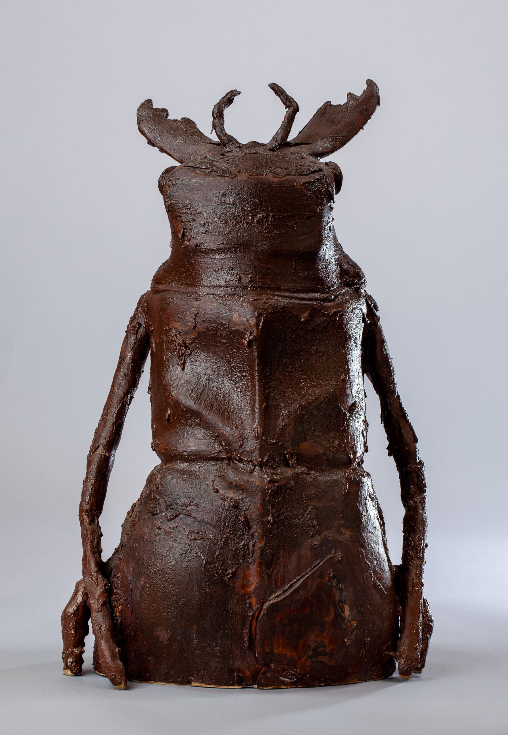 Sculpture abstraite d'un insecte ou d'une créature humanoïde, marron foncé texturé, avec des cornes et des bras pendants.