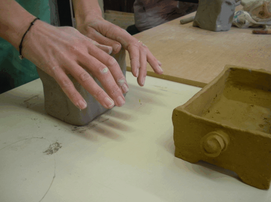 Poterie artisanale : Façonner un bloc d'argile brute. Mains façonnant un grand bloc d'argile grise dans un atelier. Une boîte en terre cuite brun-ocre est posée sur la table blanche.